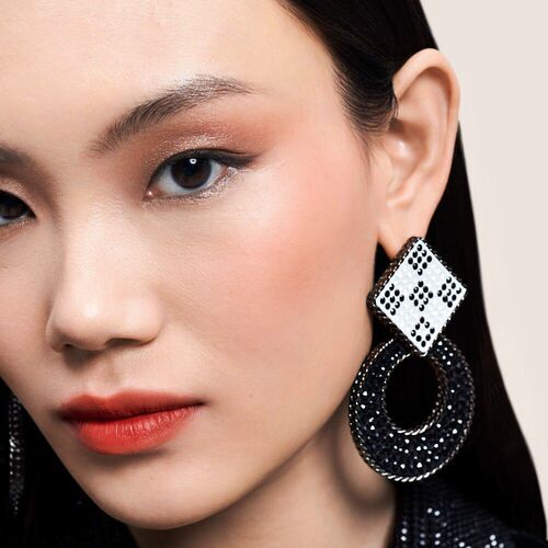 Mujer con maquillaje elegante y pendientes grandes, uno negro y dorado con un diseno cuadrado blanco y negro.
