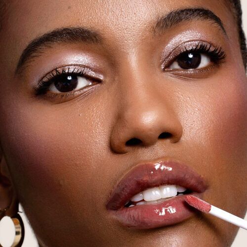 A close-up of a woman applying lip gloss, featuring glowing skin and shimmering eyeshadow.