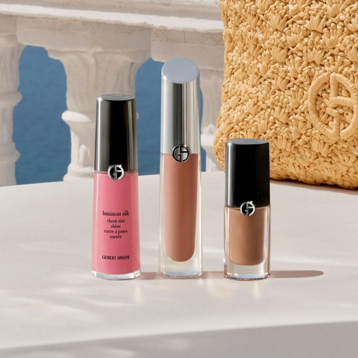 Productos de maquillaje Giorgio Armani expuestos sobre un pedestal blanco: un colorete liquido rosa, un brillo de labios nude y un pintalabios liquido marron, en un entorno de inspiracion mediterranea con arcos de piedra, cielo azul y un accesorio dorado texturizado.