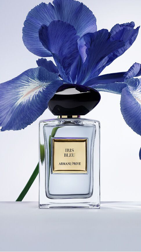 Armani/Prive Iris Bleu: un frasco de cristal transparente con tapon de piedra negra, frente a una flor de iris azul vibrante, capturando un equilibrio delicado entre elegancia y profundidad.