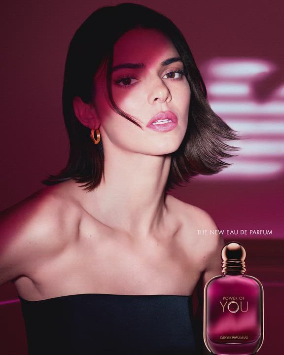 Un anuncio del Eau de Parfum "Power of You" de Emporio Armani. La imagen muestra a la modelo Kendall Jenner a la izquierda, mirando hacia la camara con una expresion suave. Tiene el cabello corto y oscuro, y viste un top negro sin tirantes y pendientes de aro dorados. A la derecha, se muestra un frasco de perfume de color purpura rosado intenso con las palabras "Power of You" y "Emporio Armani". Al fondo, aparece el logotipo del aguila de Armani, estilizado y brillante, sobre un fondo magenta oscuro. El texto dice "POWER OF YOU" arriba a la derecha y "THE NEW EAU DE PARFUM" abajo a la derecha.
