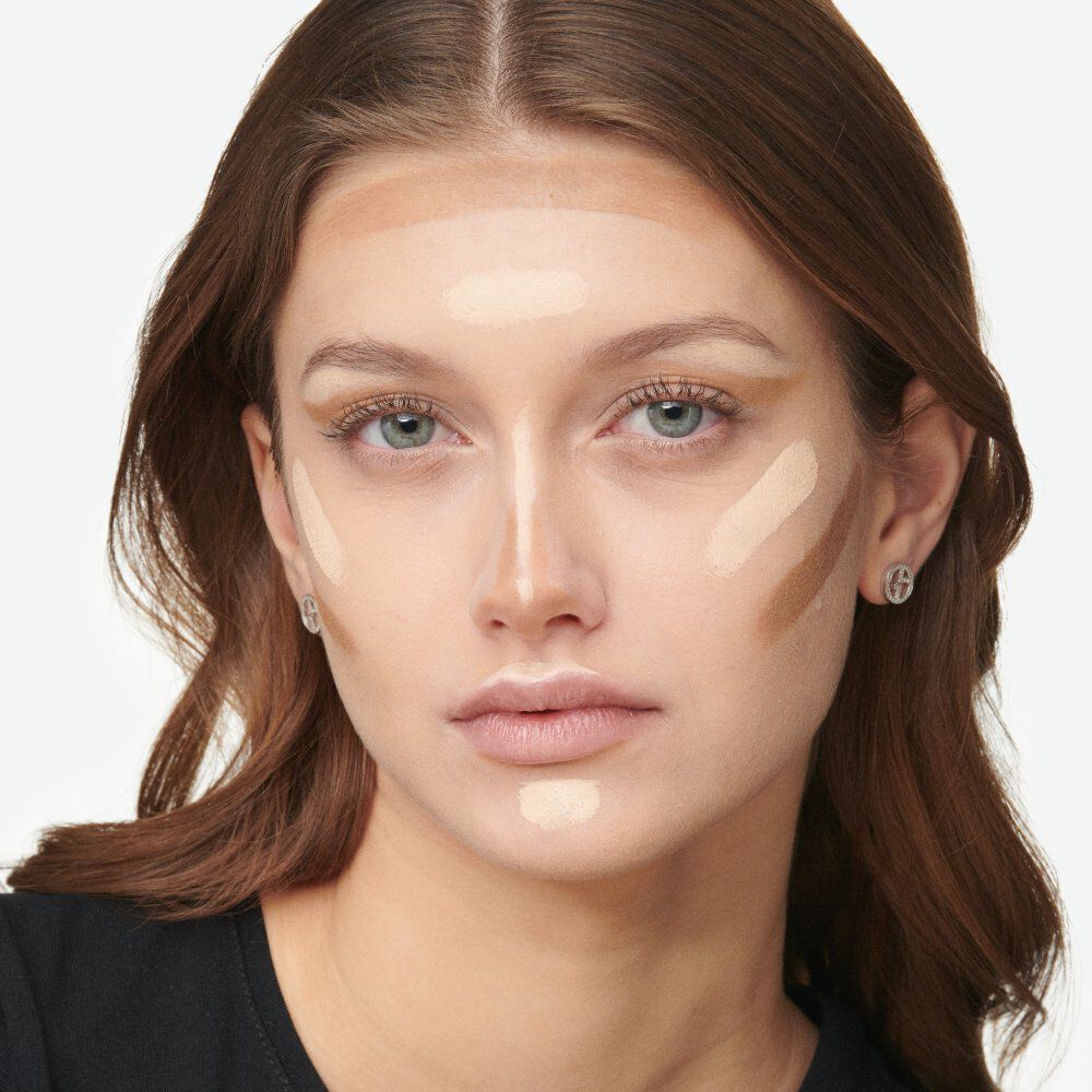 A close-up of a woman applying makeup, featuring contour and highlight shades on her face. She has a neutral expression and her hair is styled loosely. The background is plain and light-colored.