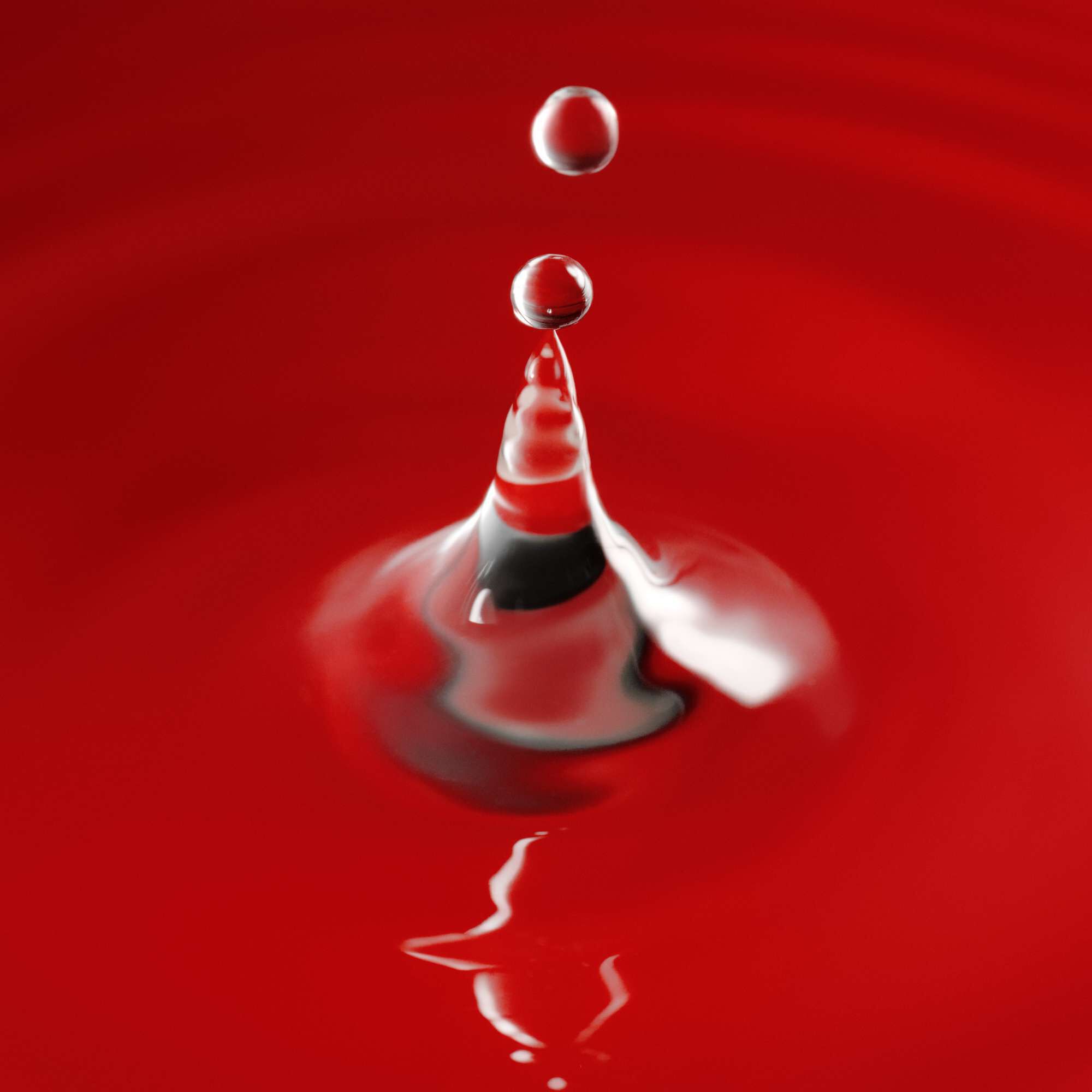A close-up of a water droplet creating ripples on a red surface, with a blurred reflection beneath it.
