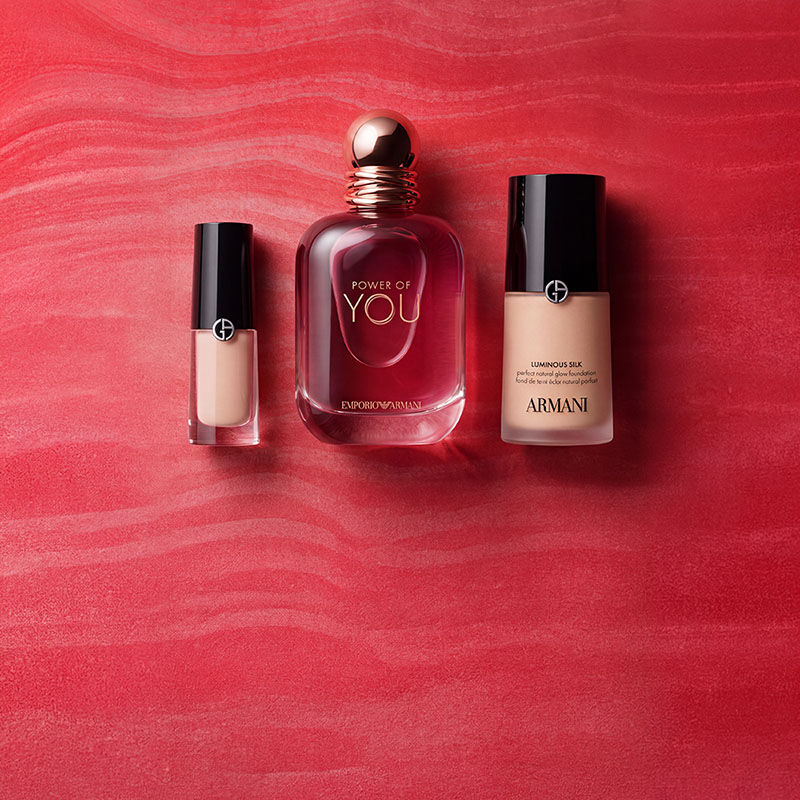 Un neceser rojo de Armani Beauty rodeado de productos de fragancias, maquillaje y cuidado de la piel sobre un fondo rojo texturizado.
