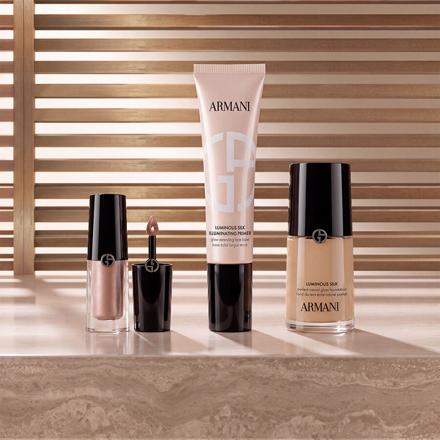 Productos de maquillaje de Armani beauty, incluyendo iluminador liquido, la prebase iluminadora Luminous Silk y la base de maquillaje Luminous Silk, dispuestos sobre una superficie de marmol con un fondo calido de listones.