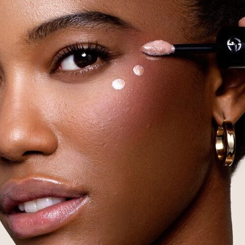 A close-up of a woman applying a shimmering product to her cheek, highlighting her radiant skin and expressive features. She is wearing gold hoop earrings and has a confident smile.