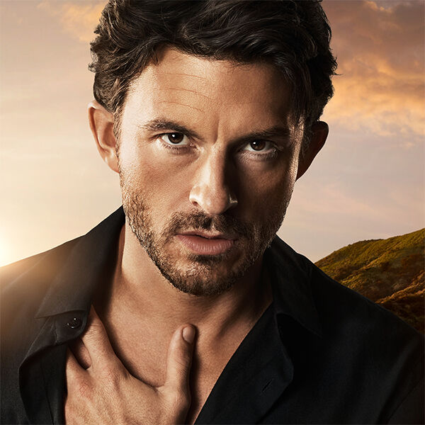 Retrato de cerca de Jonathan Bailey, cabello castano y barba corta, mirada intensa, sobre un fondo de cielo al atardecer