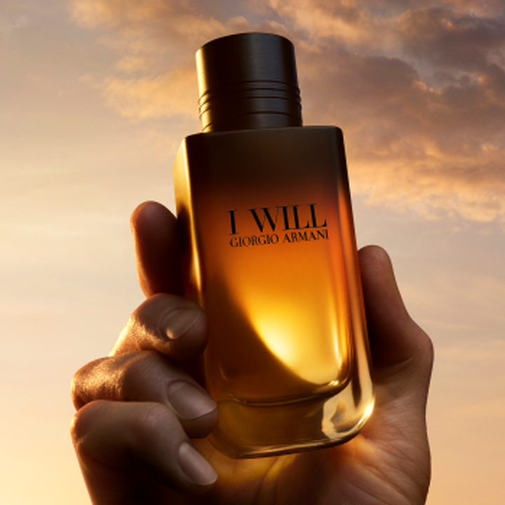 Una mano sostiene el frasco de perfume I WILL Giorgio Armani frente a un cielo dorado al atardecer.
