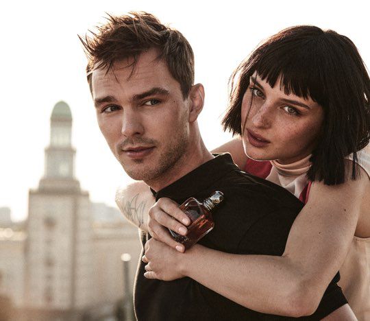 A man with short hair and a serious expression stands in the foreground, while a woman with a bob hairstyle playfully embraces him from behind, holding a perfume bottle. The setting has a soft, warm light, suggesting a sunset or dusk ambiance.