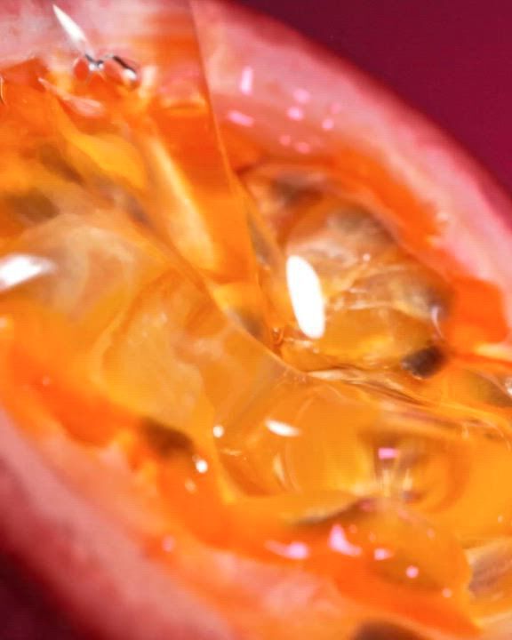 Un primer plano extremo de una maracuya (fruta de la pasion) cortada, que muestra las semillas jugosas de un naranja vibrante y la pulpa con gran detalle. La imagen tiene una textura calida y brillante. Superpuesto en texto blanco esta la descripcion: "Una explosion acida y jugosa de Maracuya (Fruta de la Pasion) con el deleite sensual y calido de la Vainilla". En la parte inferior, hay un enlace "DESCUBRIR" y un icono de reproduccion blanco.