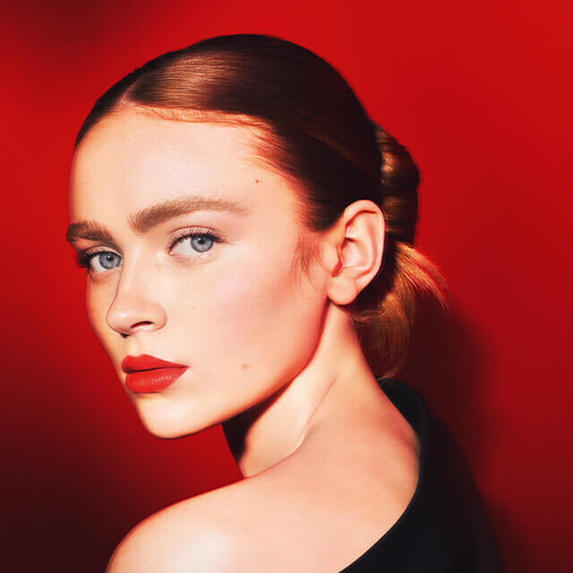 Mujer con piel clara y ojos azules, mirando hacia atras sobre un fondo rojo vibrante. Lleva un peinado elegante y un labial rojo.
