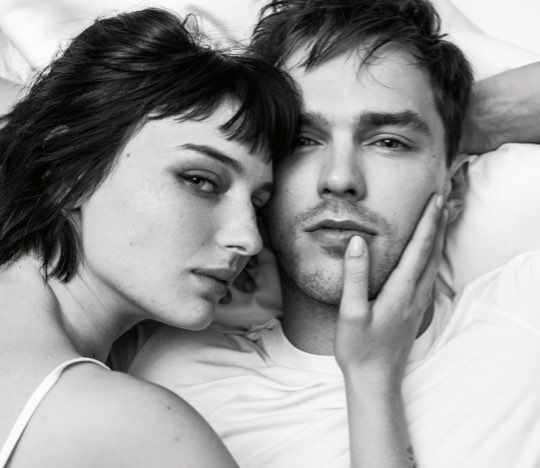 A black-and-white photo of a woman leaning her head on a man's shoulder, with her hand on his face. They are both looking directly at the camera with serious expressions, surrounded by soft pillows.