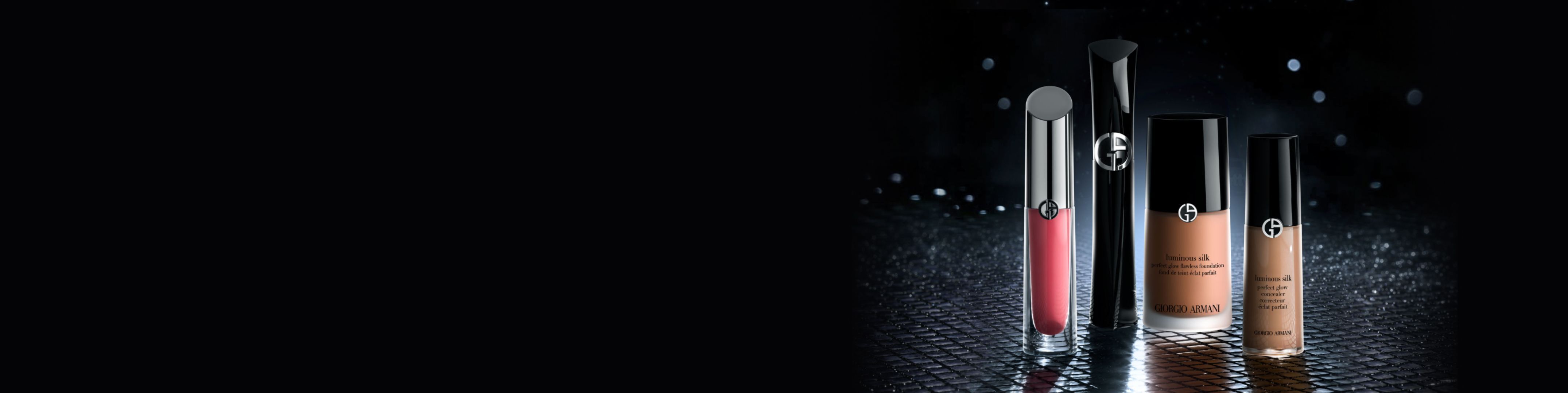 Maquillaje de Armani Beauty sobre fondo oscuro: laca de labios roja, mascara negra, base Luminous Silk y corrector sobre superficie reflectante.