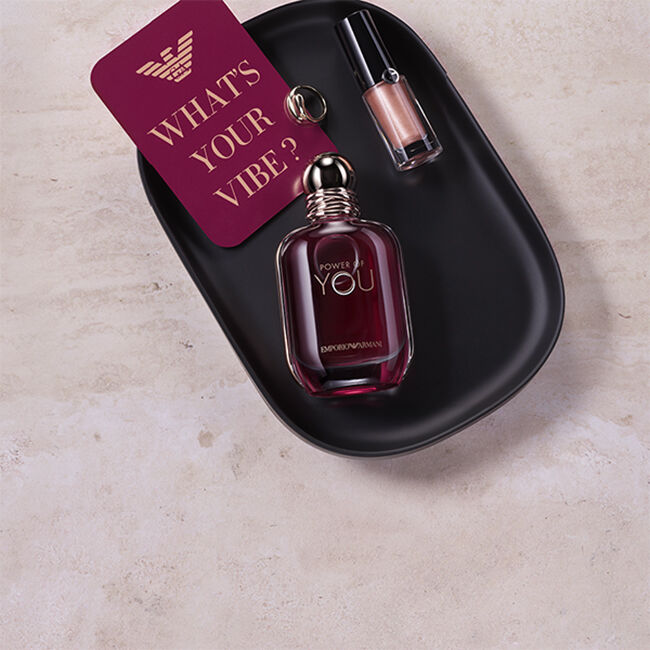 Frasco de la fragancia Power of You de Emporio Armani y producto de labios metalizado sobre una bandeja negra, junto a una tarjeta con el mensaje « What’s Your Vibe? ».