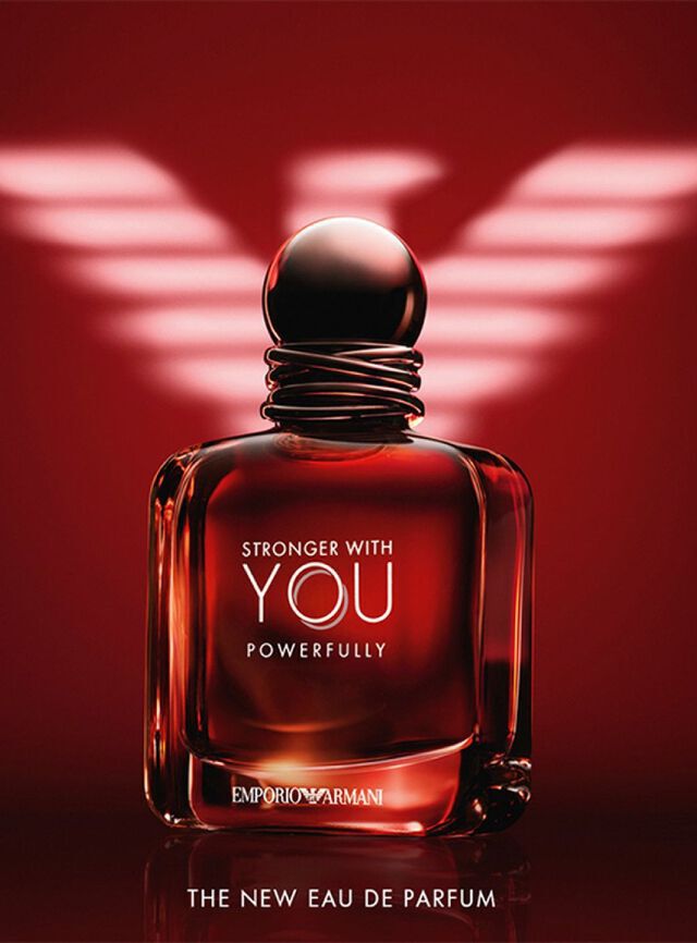 Un anuncio impactante del Eau de Parfum "Stronger With You Powerfully" de Emporio Armani. La imagen muestra un frasco de vidrio cuadrado y robusto, lleno de un liquido rojo ambarino intenso que brilla desde el interior. El frasco tiene un tapon oscuro y redondeado con un detalle de hilos entrelazados alrededor del cuello. Las etiquetas "STRONGER WITH YOU POWERFULLY" y "EMPORIO ARMANI" estan impresas en plata y blanco. Al fondo, un gran logotipo del aguila de Armani estilizado esta iluminado en blanco sobre un fondo rojo profundo. En la parte inferior, el texto dice "THE NEW EAU DE PARFUM".