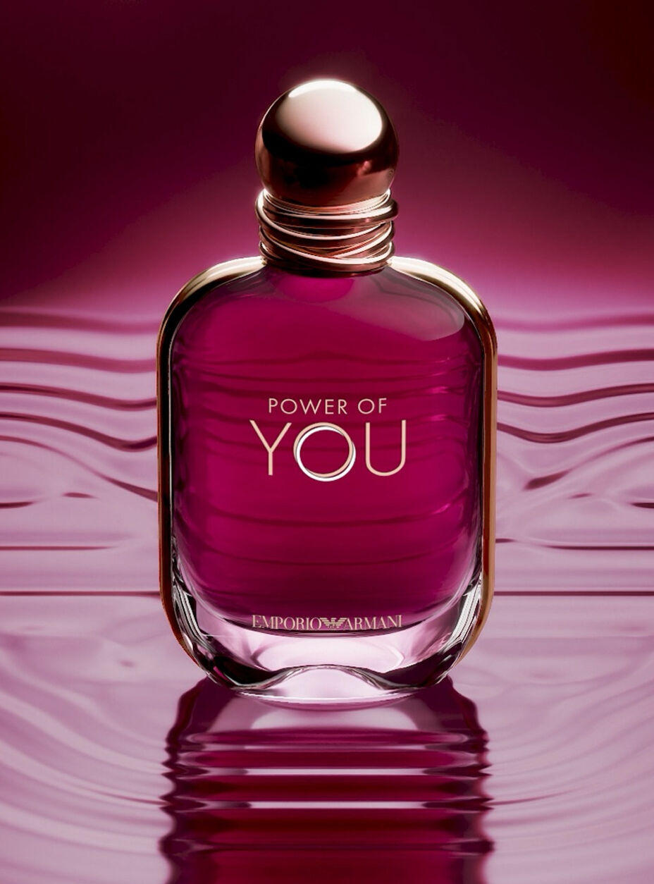  Un anuncio en primer plano del frasco de perfume "Power of You" de Emporio Armani. El frasco de vidrio en tonos rubi se encuentra en el centro sobre una superficie liquida reflectante de color magenta oscuro que muestra suaves ondas. El frasco tiene un tapon dorado redondeado y las palabras "POWER OF YOU" y "EMPORIO ARMANI" impresas en oro. El fondo es un degradado suave y desenfocado de rosa profundo y purpura. En la parte inferior, un texto blanco dice: "Presentamos un nuevo frasco iconico: una joya en tonos rubi", seguido de un enlace "DESCUBRIR" y un icono de pausa.