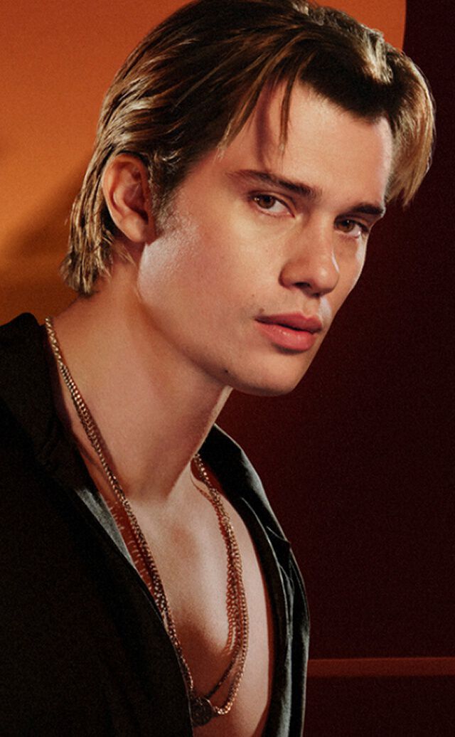 Un anuncio de la gama de fragancias "Stronger With You" de Emporio Armani. A la izquierda, un joven de cabello castano claro viste una camisa negra y collares de plata, mirando hacia la camara con una expresion seria. A la derecha, un carrusel de productos bajo el titulo "EVERY FACET OF STRONGER WITH YOU" muestra tres frascos de fragancia diferentes. El frasco central resaltado es "Stronger With You Powerfully" en tamano de 50 ml, con un precio de £74.00, y un boton de "ANADIR A LA CESTA" debajo. Tambien se ven flechas de navegacion y una barra deslizante.