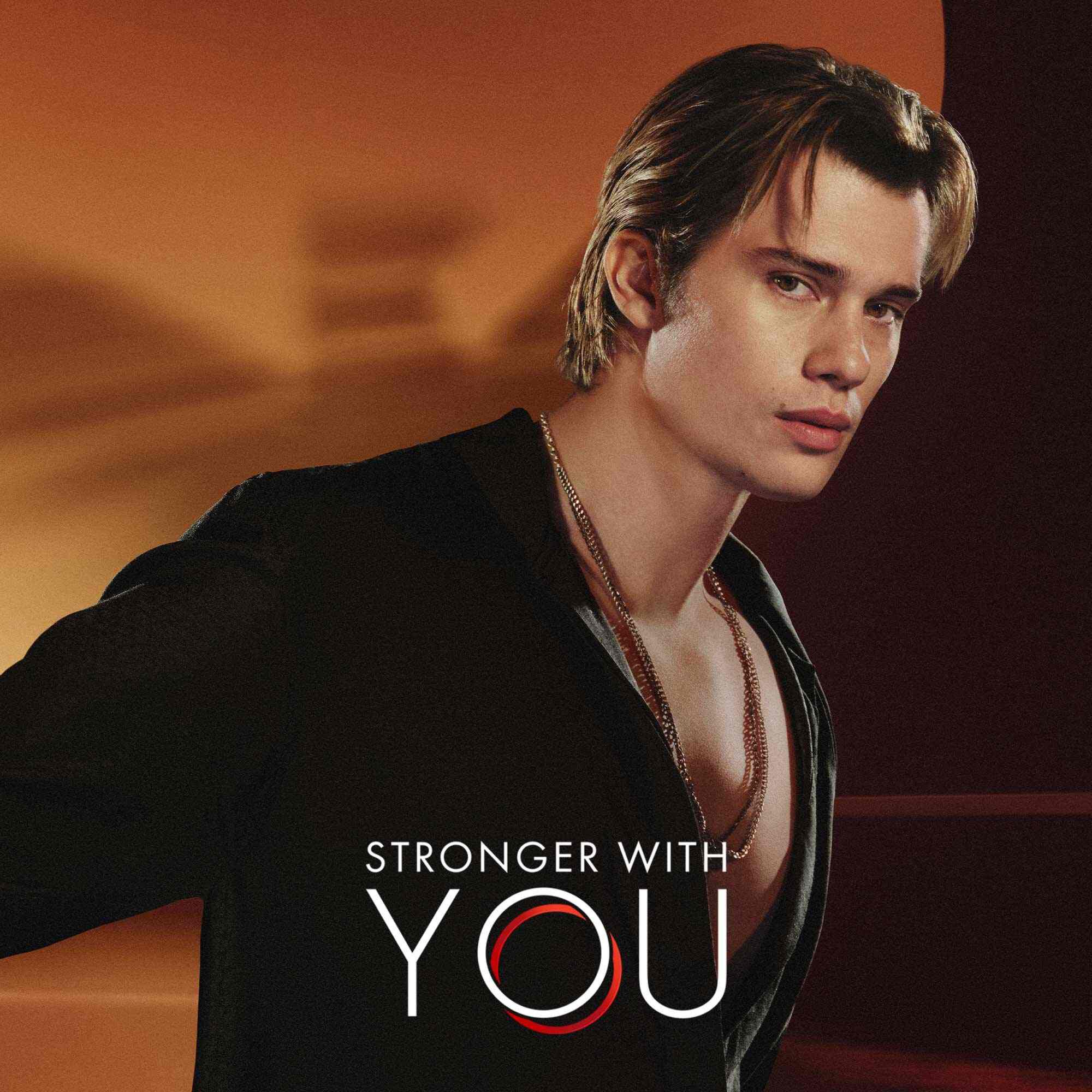 Nicholas Galatzine con camisa negra y el texto del logo 'Stronger With You' sobre un fondo naranja calido
