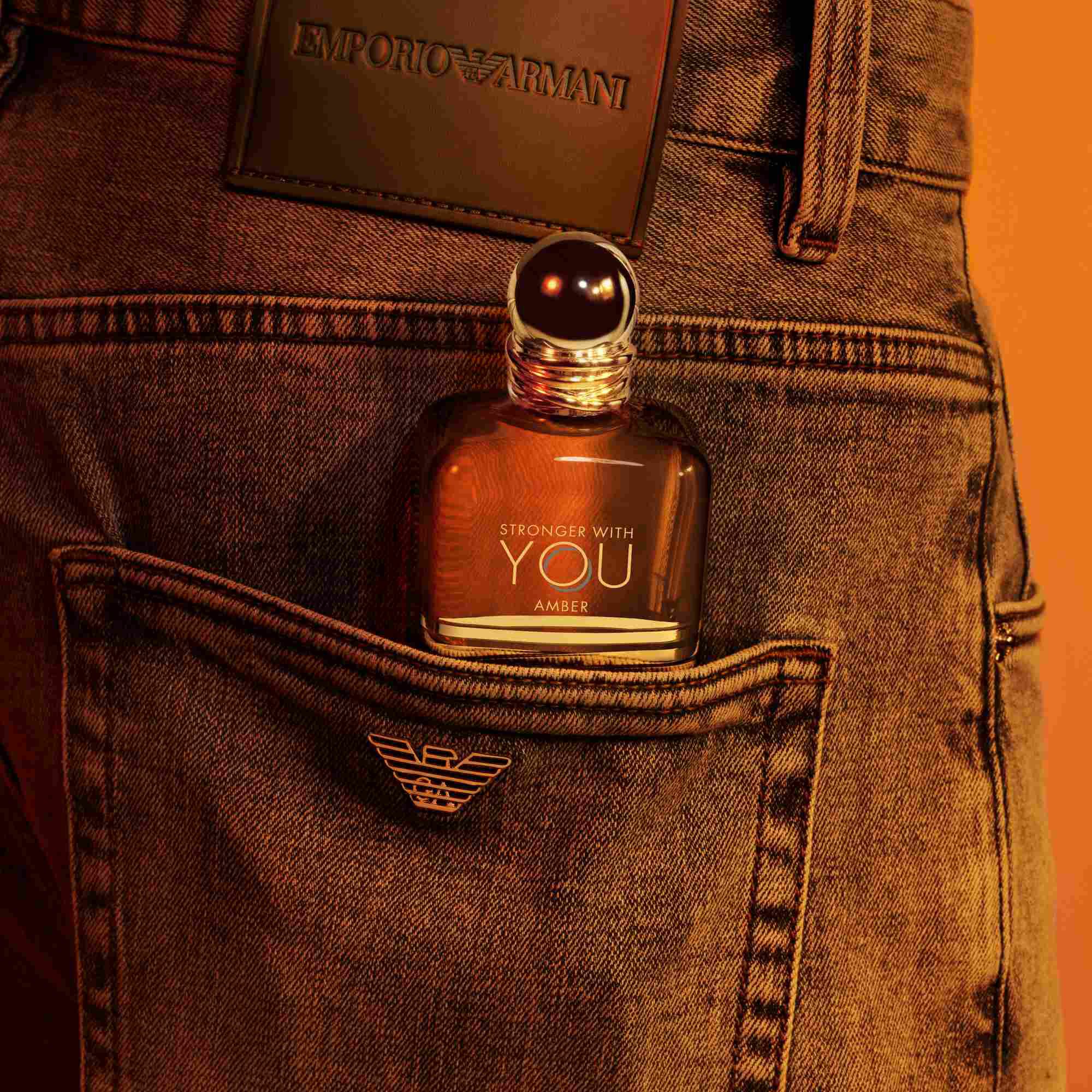 Un frasco de perfume Emporio Armani Stronger With You Amber asomandose del bolsillo trasero de unos jeans, con un parche de cuero oscuro visible por encima