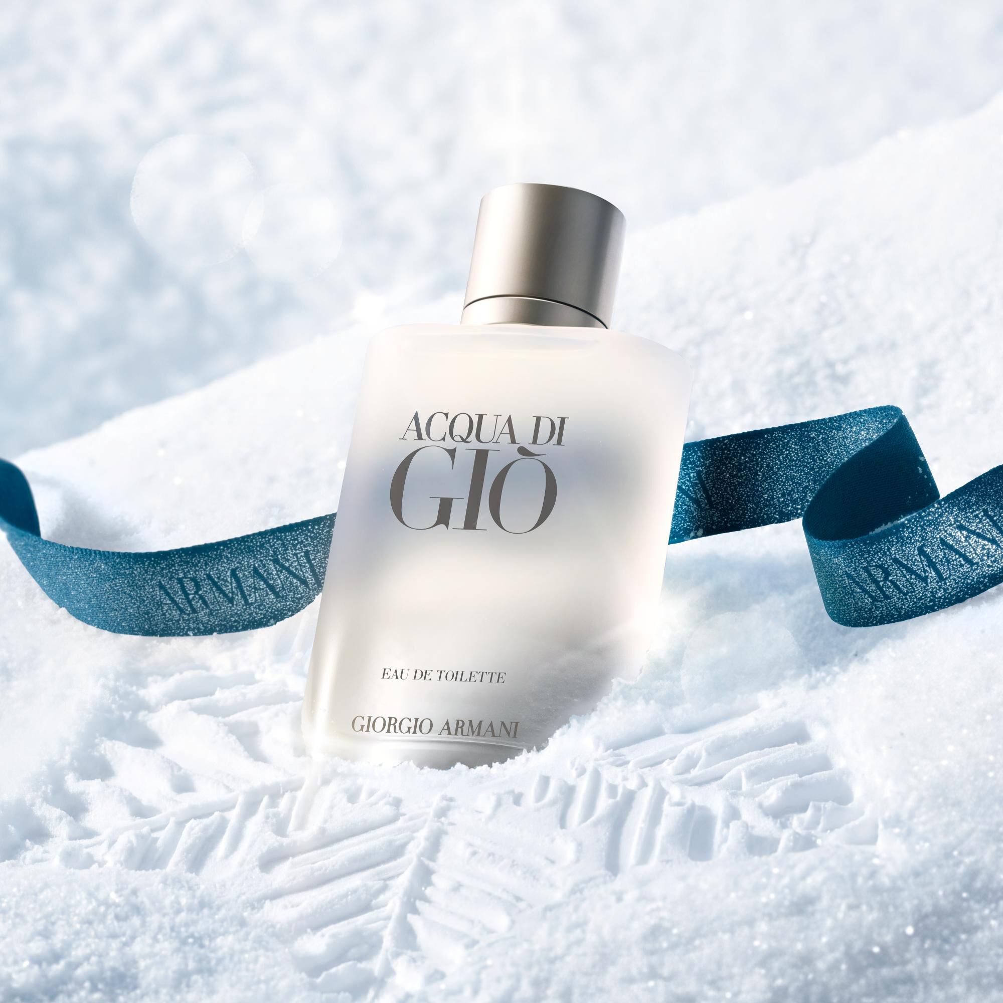 Acqua di Gio Eau de Toilette by Giorgio Armani, clear glass bottle resting in snow with a blue Armani ribbon — men's aquatic fragrance, holiday gifting visual.