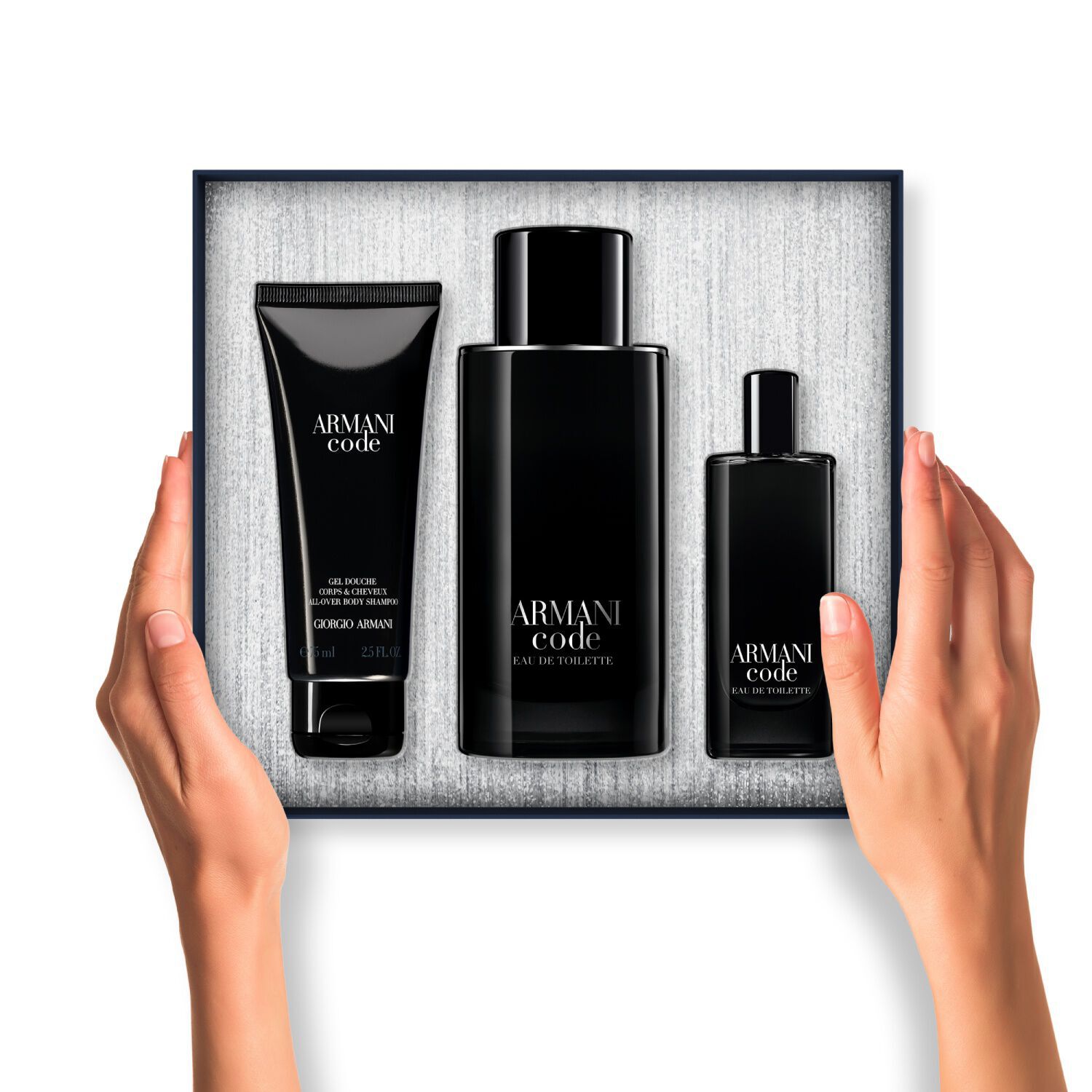 Un par de manos sostiene una caja de regalo abierta de Armani Code. Dentro, tres productos envasados en negro (gel corporal, Eau de Toilette grande y Eau de Toilette tamano viaje) estan cuidadosamente dispuestos sobre un fondo plateado texturizado.