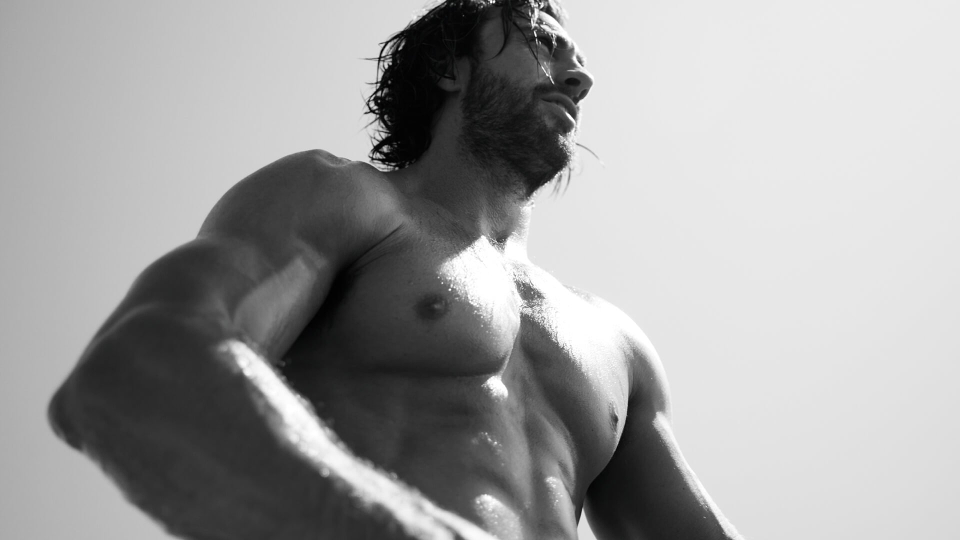 Hombre musculoso de pie, con el torso descubierto, bajo un cielo claro, mostrando una expresión de concentración.