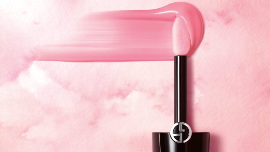 A sleek lipstick applicator with a vibrant pink gloss, set against a soft pink watercolor backdrop.