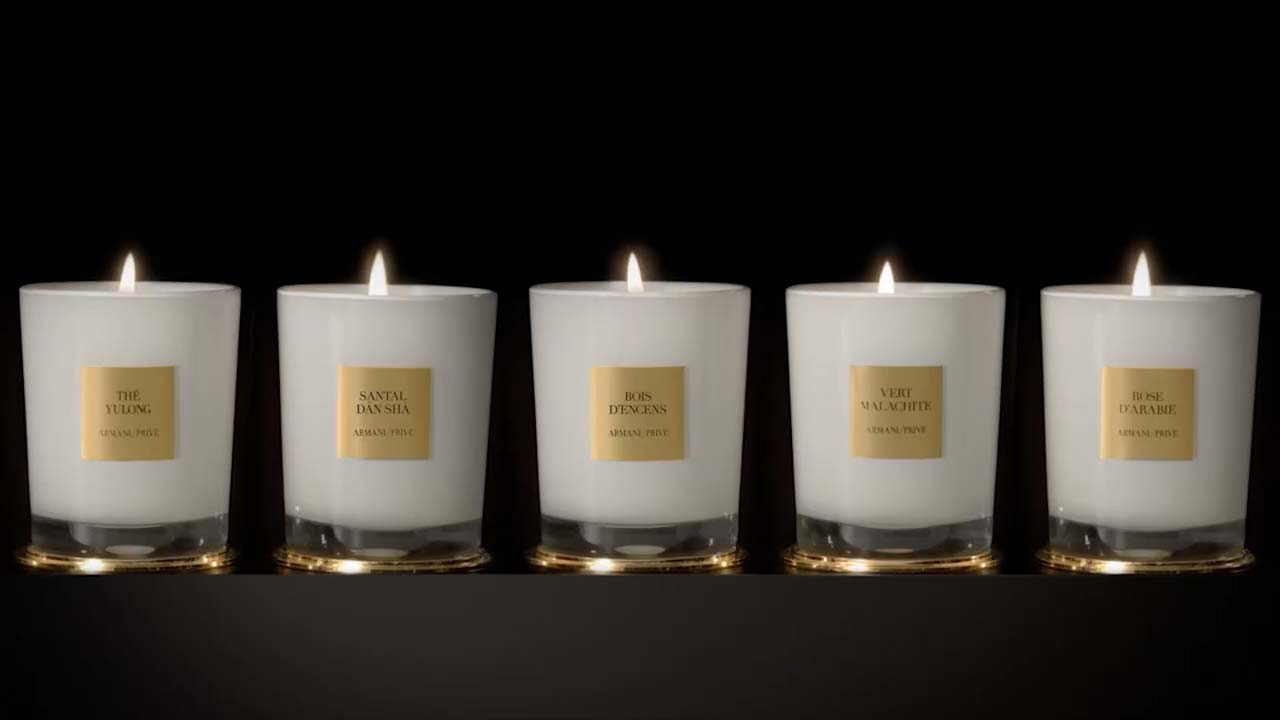 Cinco velas encendidas en frascos blancos, cada una con etiquetas doradas que indican diferentes fragancias.